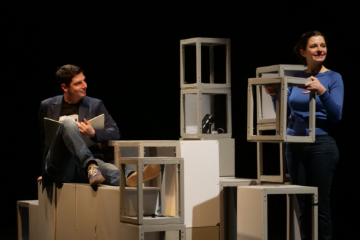 un home et une femme sur scène, l'homme est assis sur une structure en bois entourée de cubes sans fonds, la femme est debout avec l'un de ces cubes en mains - Agrandir l'image, fenêtre modale