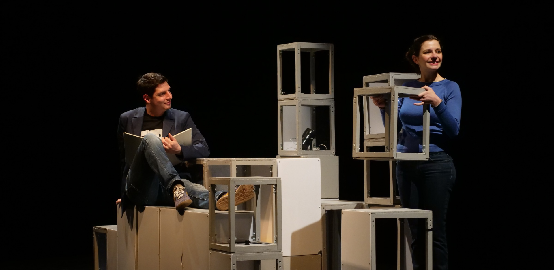 un home et une femme sur scène, l'homme est assis sur une structure en bois entourée de cubes sans fonds, la femme est debout avec l'un de ces cubes en mains
