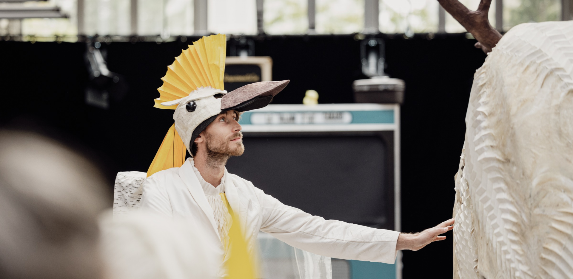 un homme en costume blanc, porte un chapeau en forme de tête d'oiseau à crête jaune